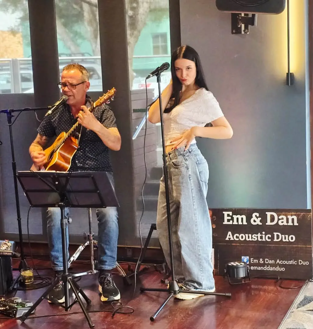 Dan playing guitar and singing while Em performs beside the Em & Dan Acoustic Duo banner