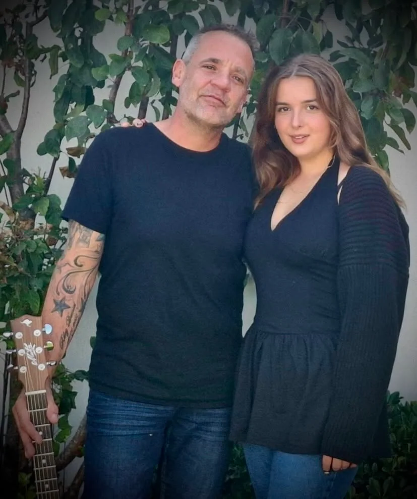 Dan and Em standing together in a garden, Dan holding an acoustic guitar