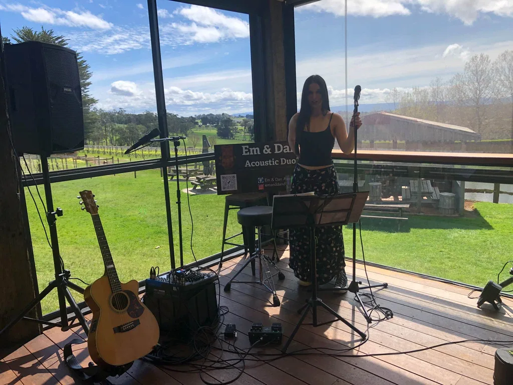 Em at the microphone on a deck overlooking a vineyard, with the duo’s guitar and sound gear
